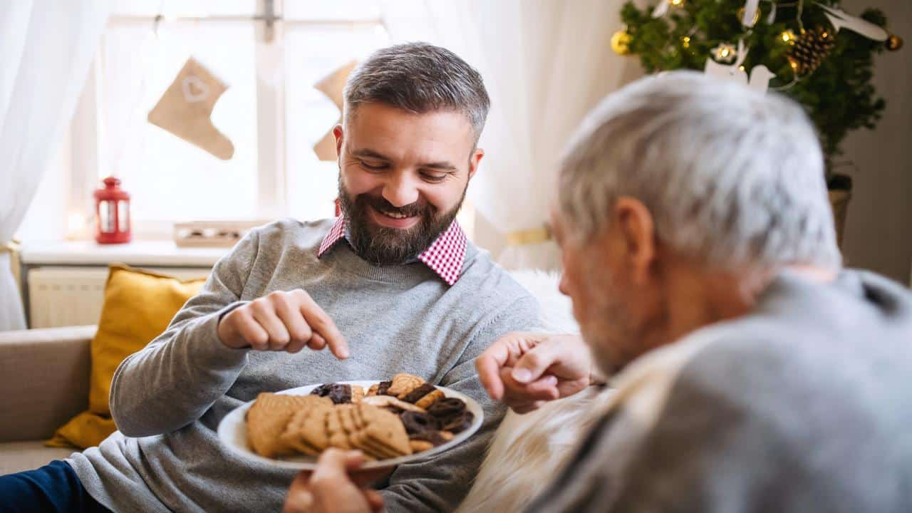 A smiling man offers a plate of assorted cookies to an older man in a cozy, festive living room.