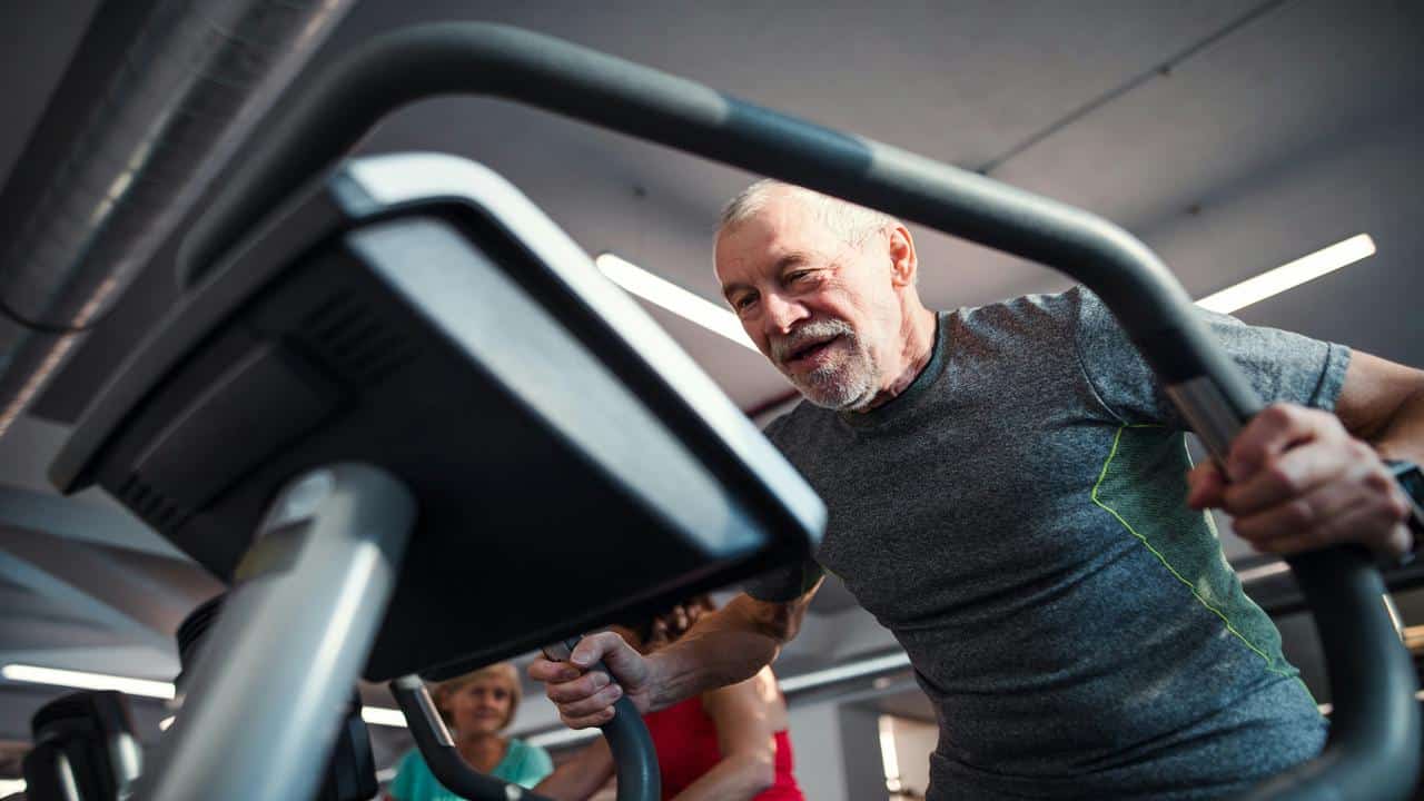 A determined older man exercises on an elliptical machine in a modern gym.