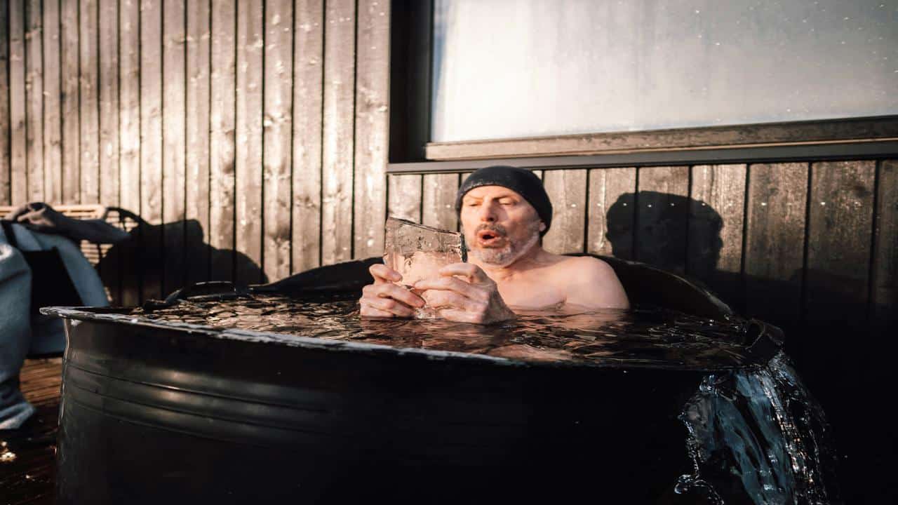 A shirtless man in a beanie sits in an overflowing ice bath, holding a large block of ice and exhaling sharply.