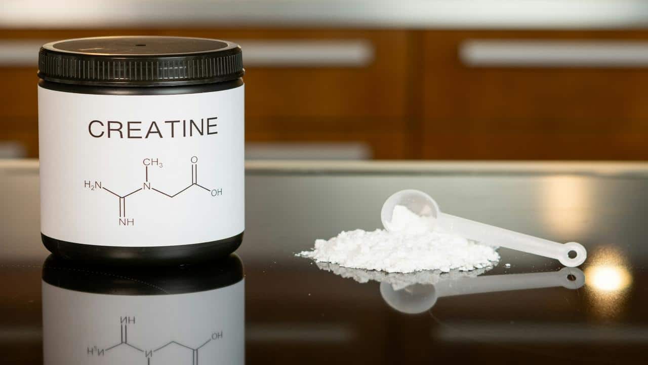 A container labeled "Creatine" with a chemical structure diagram sits beside a scoop of white powder on a reflective surface.