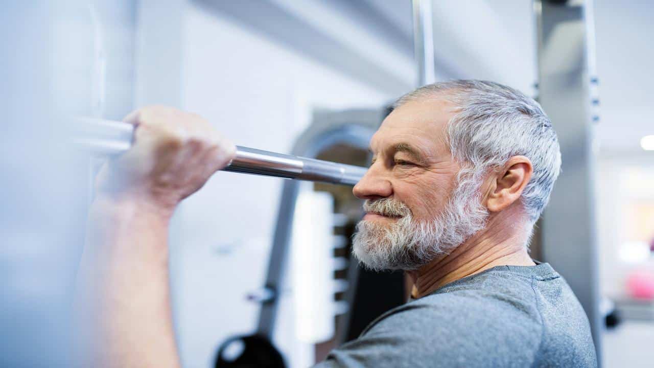 A smiling older man lifts a barbell in a gym, showing focus and strength during his workout.