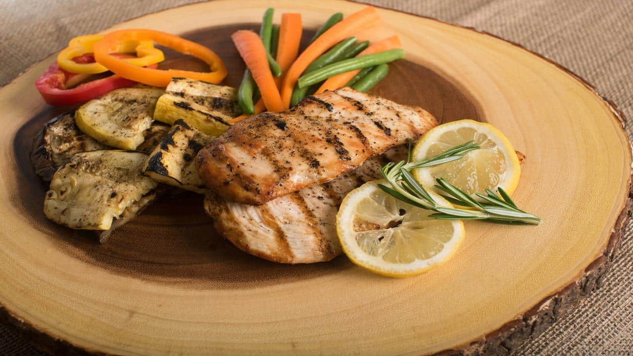 A wooden platter displays grilled chicken breast with lemon slices, rosemary, and a colorful mix of grilled and fresh vegetables.
