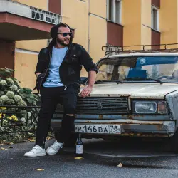 A man in casual streetwear leaning against a rusty old car in an urban setting.