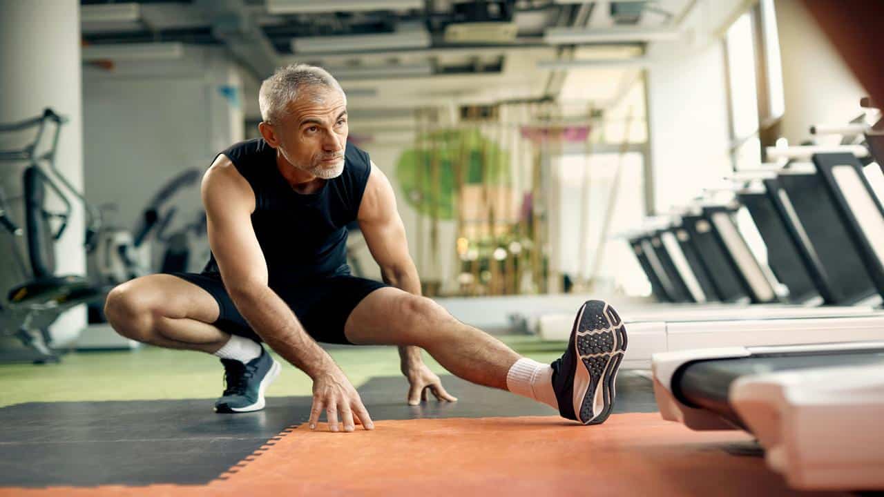 An active older man in workout clothes stretches his legs on a gym floor, preparing for exercise.
