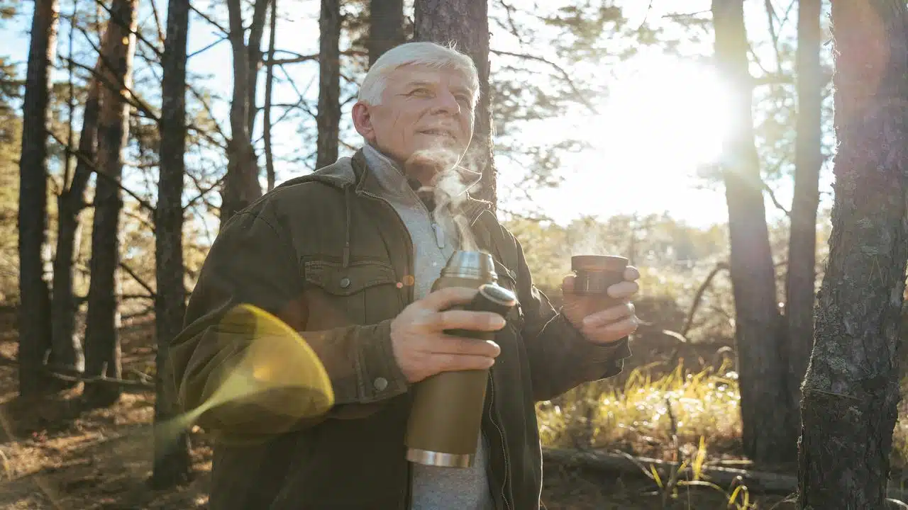 An older man holds a steaming drink in a sunlit forest, enjoying the warmth and nature around him.