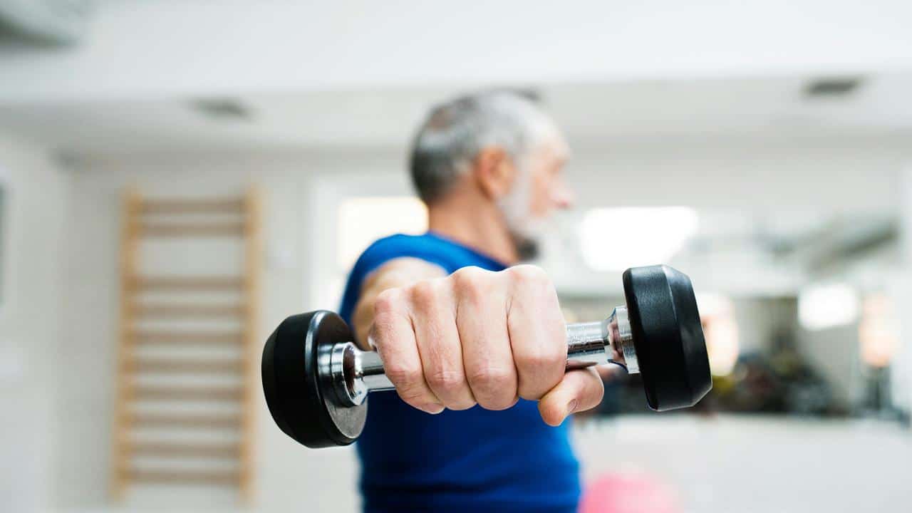A focused older man lifts a dumbbell straight ahead during a workout.
