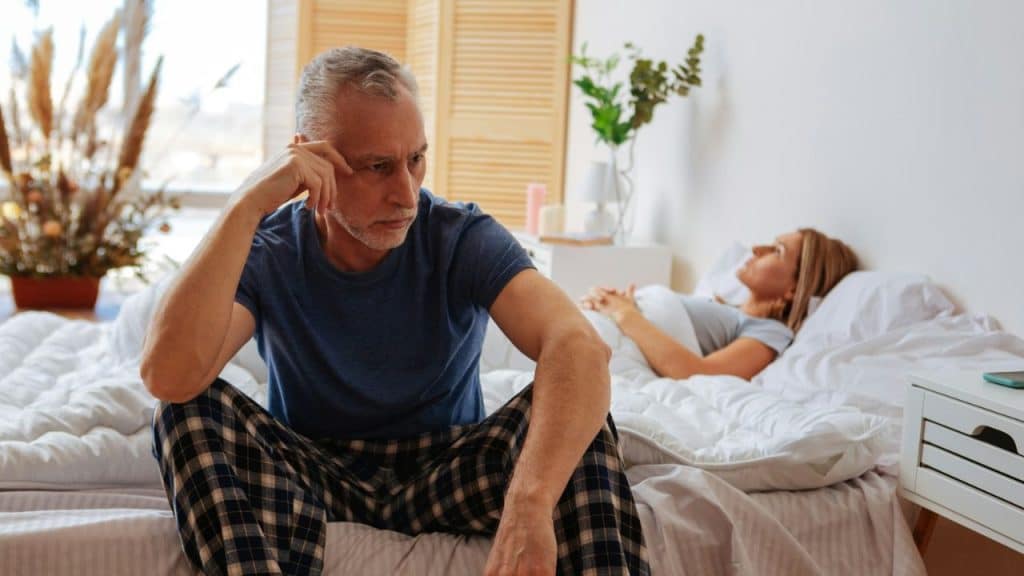 A distraught man sits on a bed, while a woman lies in the background.