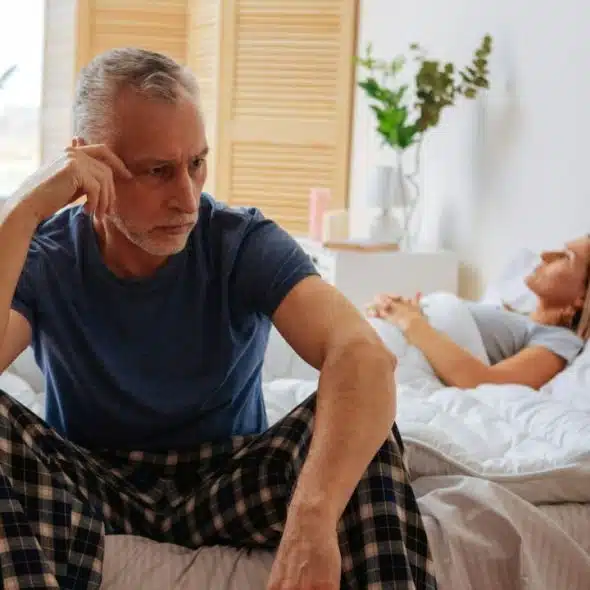 A distraught man sits on a bed, while a woman lies in the background.