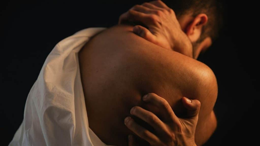 A shirtless person's back is shown as two hands clasp their shoulders, with a white cloth draped over one side.