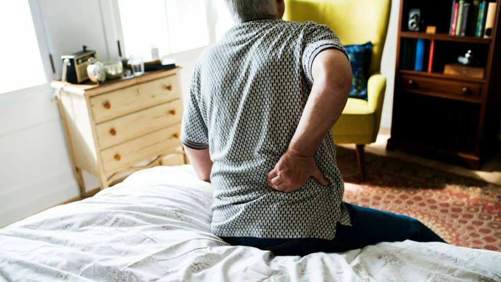 A man sits on a bed, holding his lower back in apparent pain, with a dresser and chair in the background.