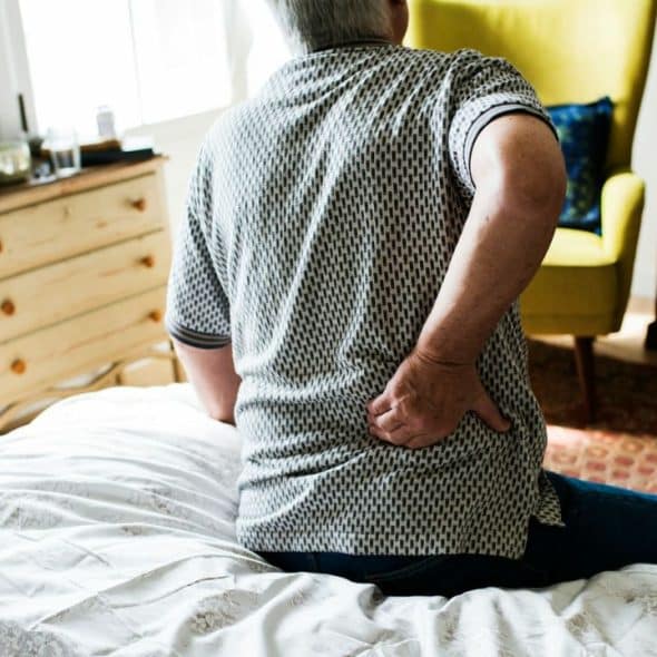 A man sits on a bed, holding his lower back in apparent pain, with a dresser and chair in the background.