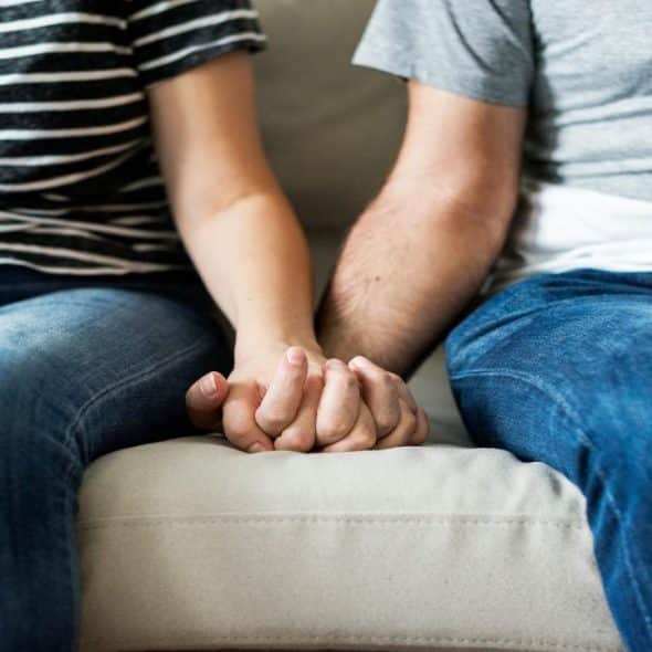 A couple sitting on a couch, holding hands, dressed casually in jeans and striped or plain t-shirts.