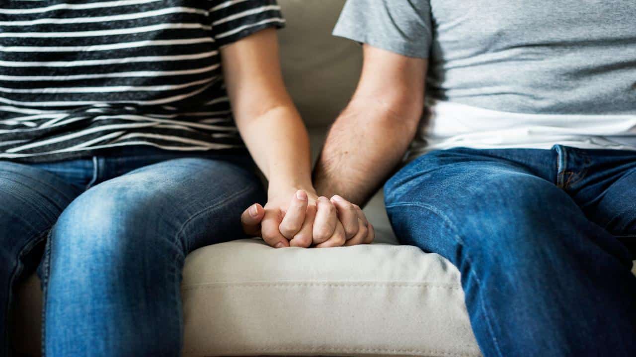 A couple sitting on a couch, holding hands, dressed casually in jeans and striped or plain t-shirts.