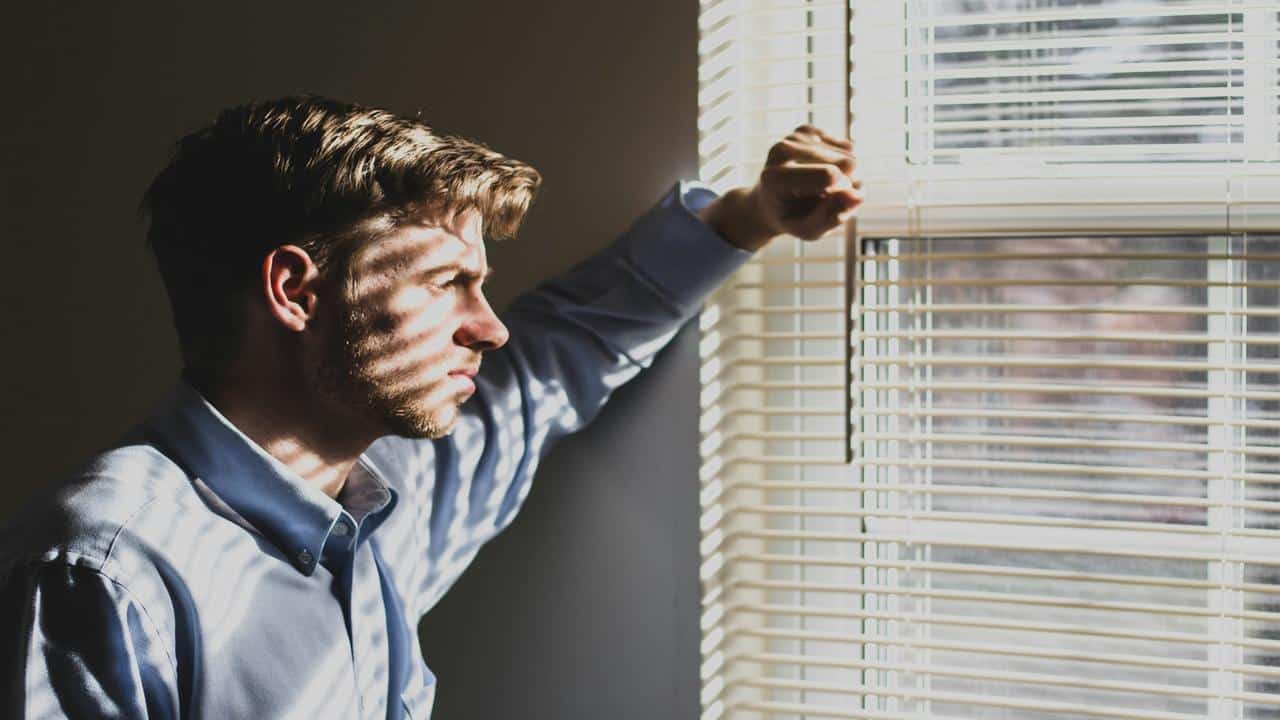 A man in a dress shirt staring pensively out a sunlit window with blinds.