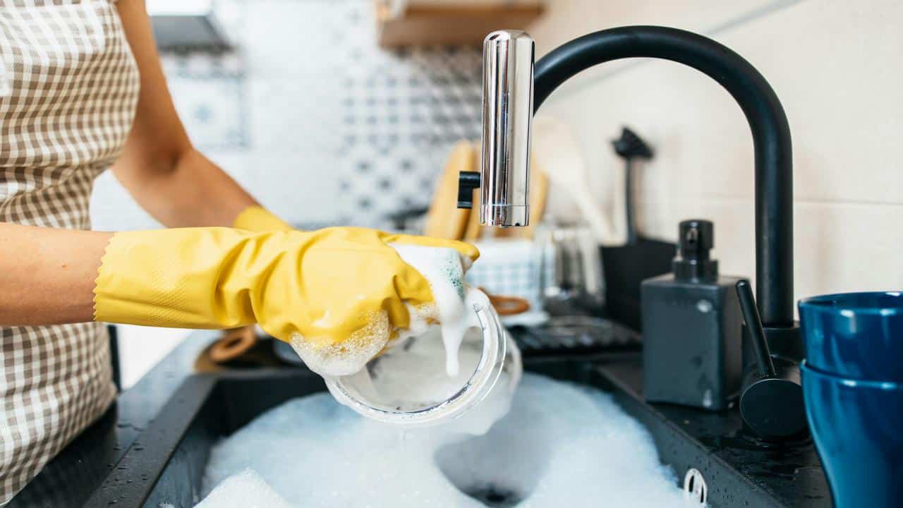 A person wearing yellow gloves washing a soapy glass in a kitchen sink.