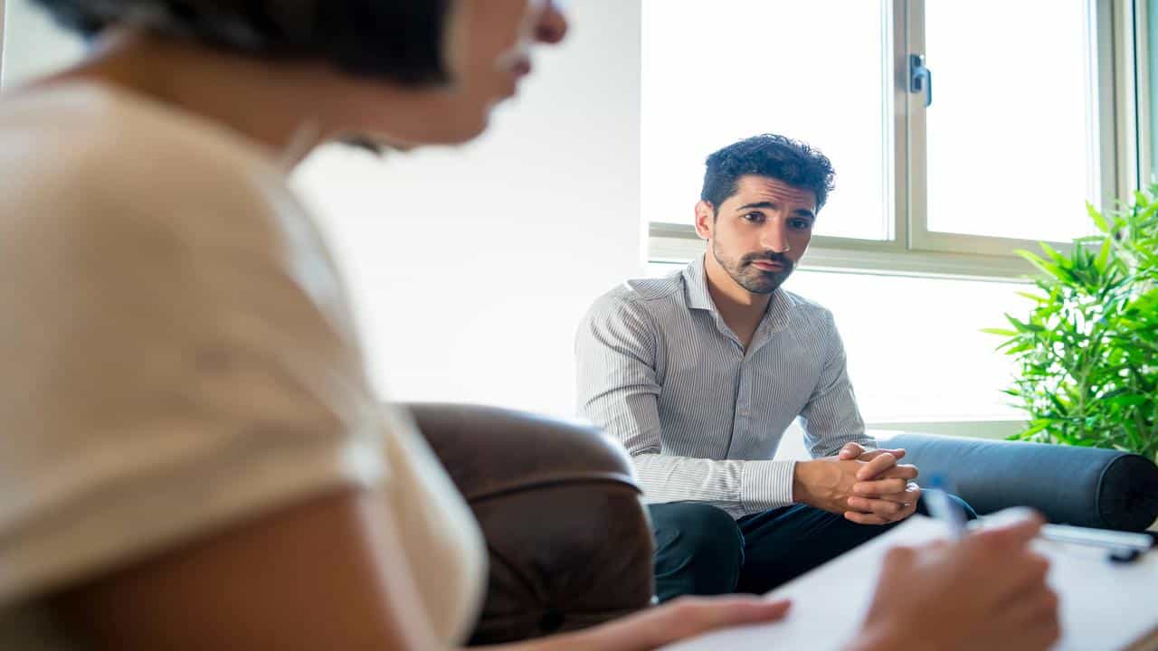A man sitting on a couch during a therapy session, looking at a counselor taking notes.