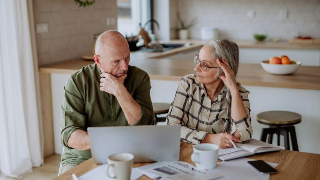 A man and woman sit at a table with a laptop, looking thoughtful.