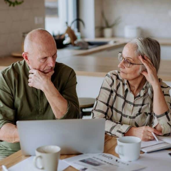 A man and woman sit at a table with a laptop, looking thoughtful.