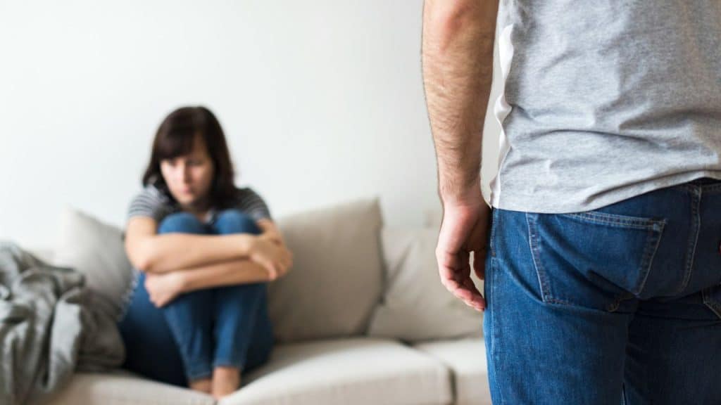 A man's back is to the camera, facing a woman sitting sadly on a sofa.
