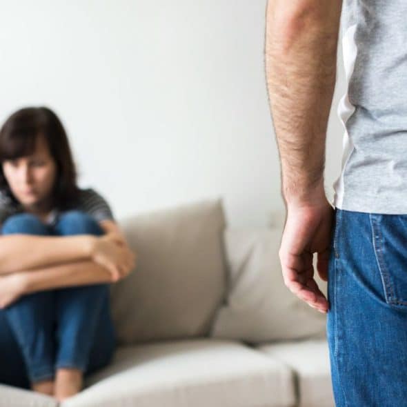 A man's back is to the camera, facing a woman sitting sadly on a sofa.