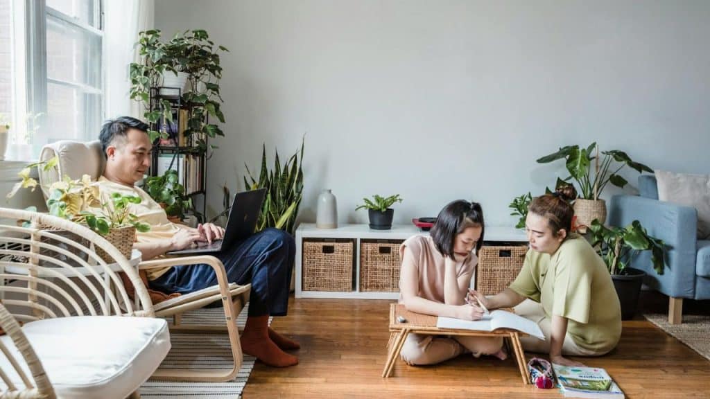 A family in a living room, with a man on a laptop and two women studying on the floor.