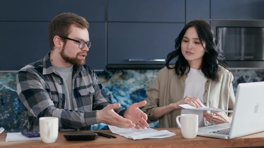 A man with glasses talks to a woman holding papers at a table with a laptop.