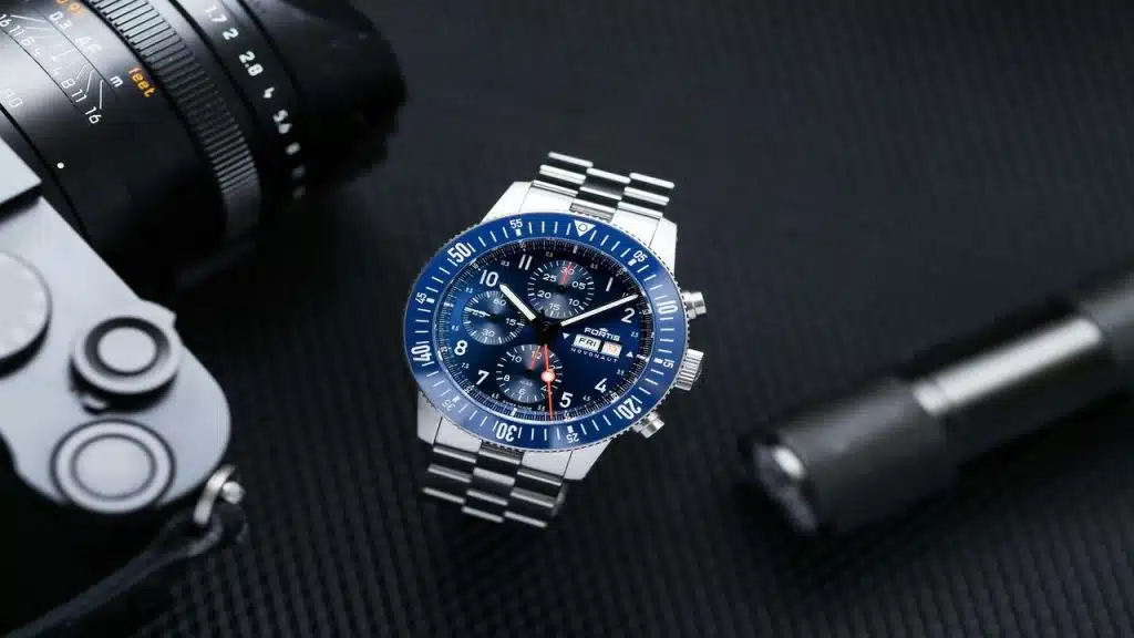 A stainless steel chronograph watch with a blue dial and bezel, placed beside a camera and flashlight.