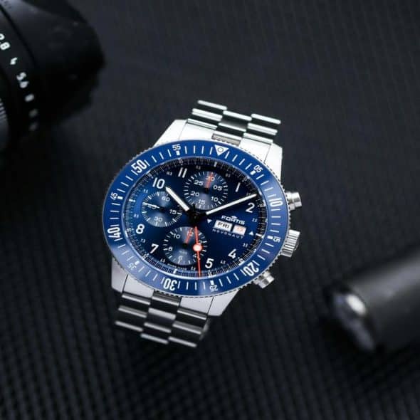 A stainless steel chronograph watch with a blue dial and bezel, placed beside a camera and flashlight.
