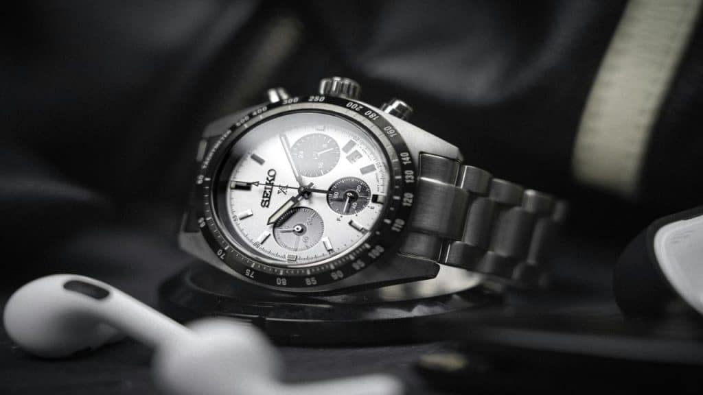 A Seiko chronograph watch with a silver dial and metal bracelet, placed near wireless earbuds.