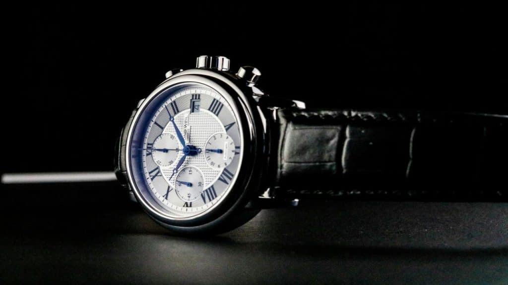 A Raymond Weil chronograph watch with a silver dial, Roman numerals, and a black leather strap, set against a dark background.