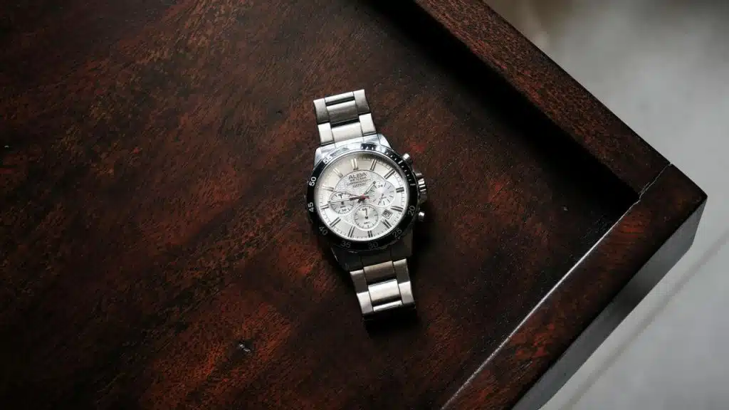 A silver Alba chronograph watch with a white dial resting on a dark wooden surface.