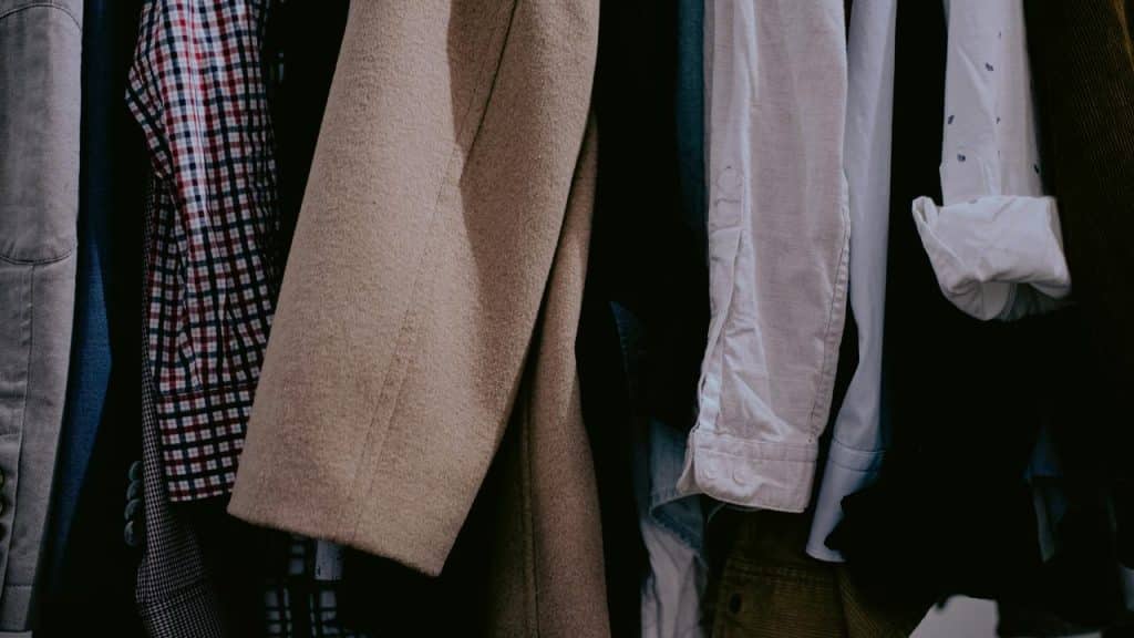 A row of men's clothing hanging in a closet, including shirts, jackets, and a beige overcoat.