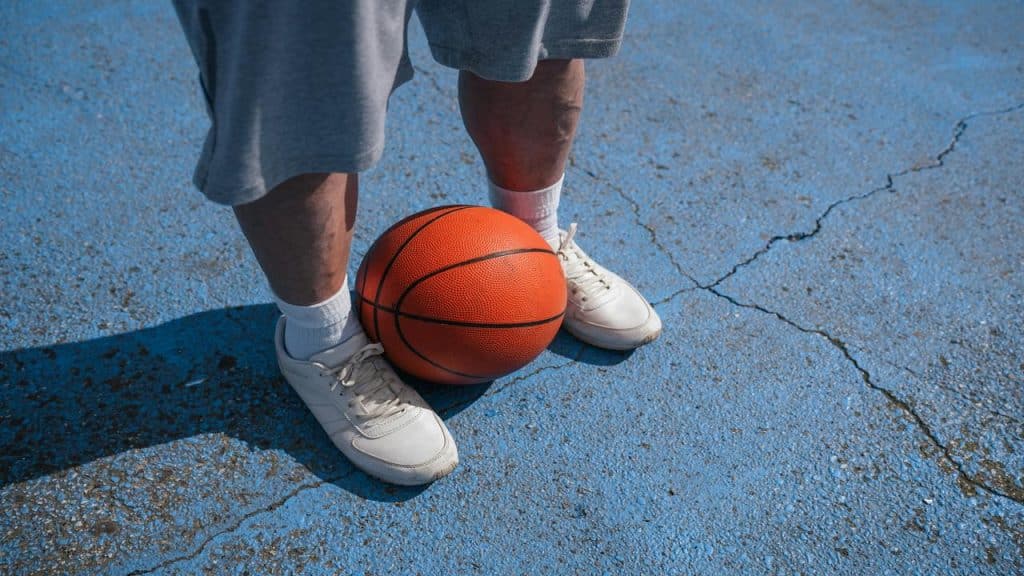 A person in white sneakers and gray shorts standing with a basketball on a blue court.
