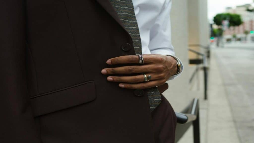 A man in a dark suit and tie adjusting his jacket, wearing rings and a watch.