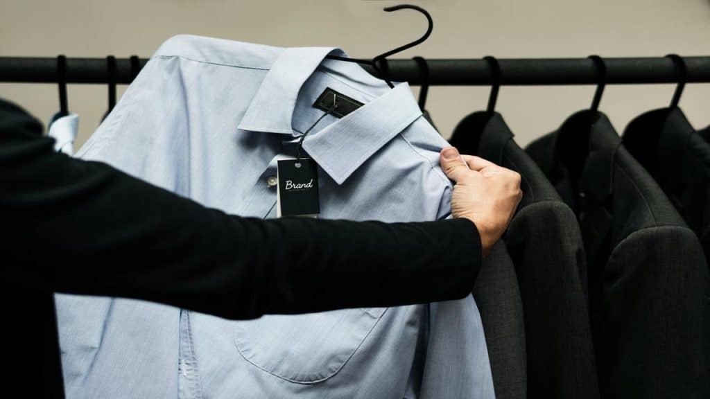 A person holding a light blue dress shirt with a tag, among dark suits on a rack.