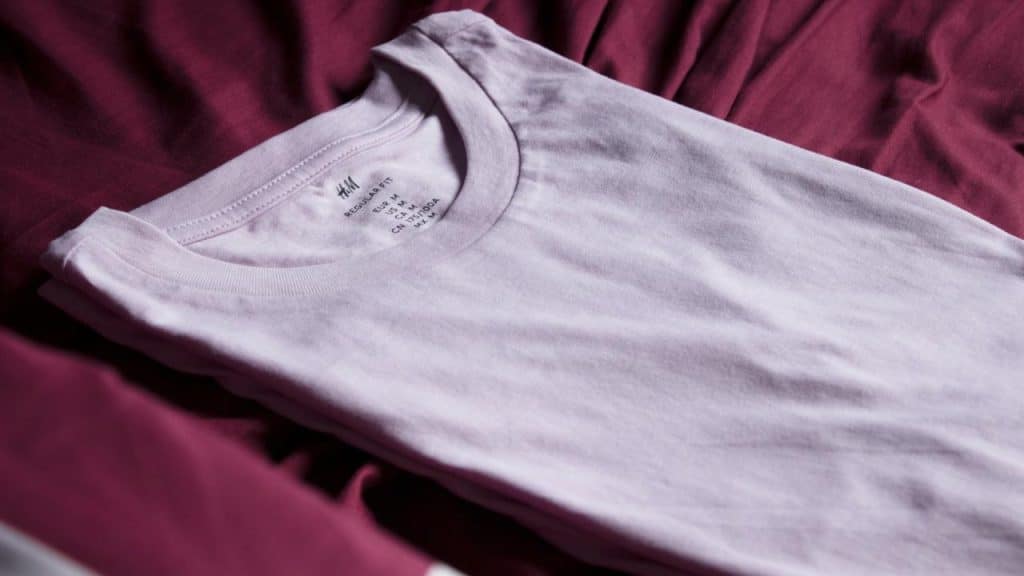 A folded light pink T-shirt placed on a maroon fabric surface.
