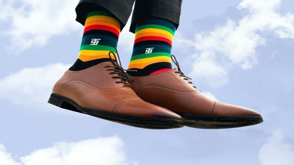 A pair of brown dress shoes worn with colorful striped socks against a sky background.