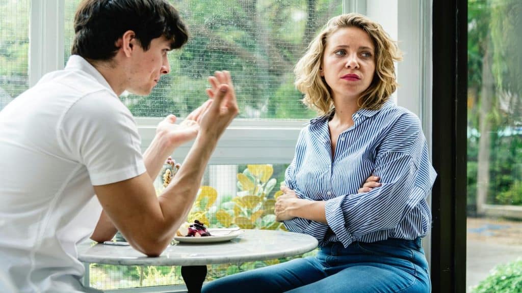 A woman with crossed arms looks upset while a man gestures during a tense conversation at a café.