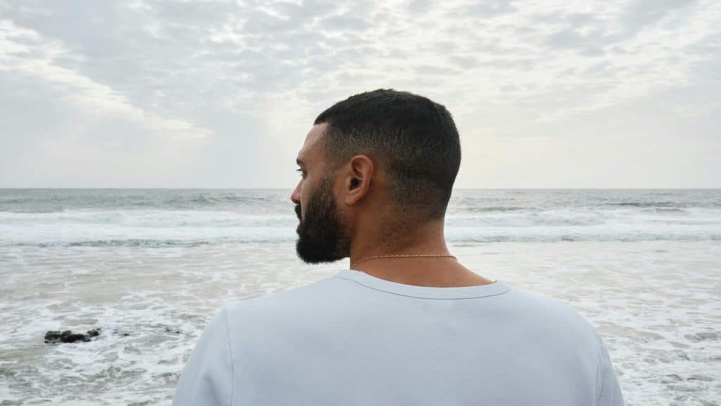 A man with a trimmed beard looks out at the ocean, standing on a beach under a cloudy sky.