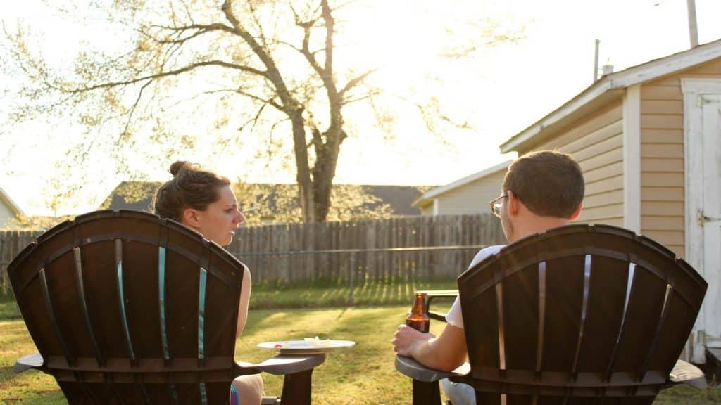 A man and woman sit in Adirondack chairs in a backyard, talking during sunset with a drink and snacks nearby.