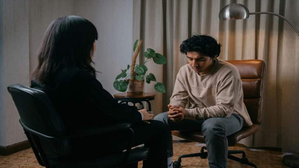 A man sits with his head down and hands clasped during a serious conversation with a therapist.