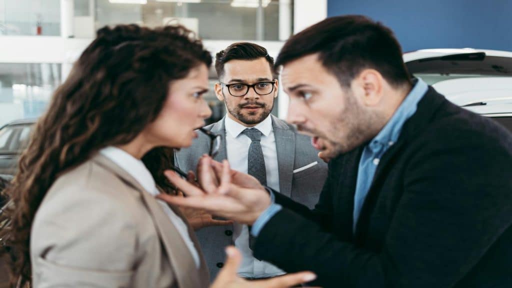 A man and woman argue intensely at a car dealership while a salesman watches uncomfortably in the background.