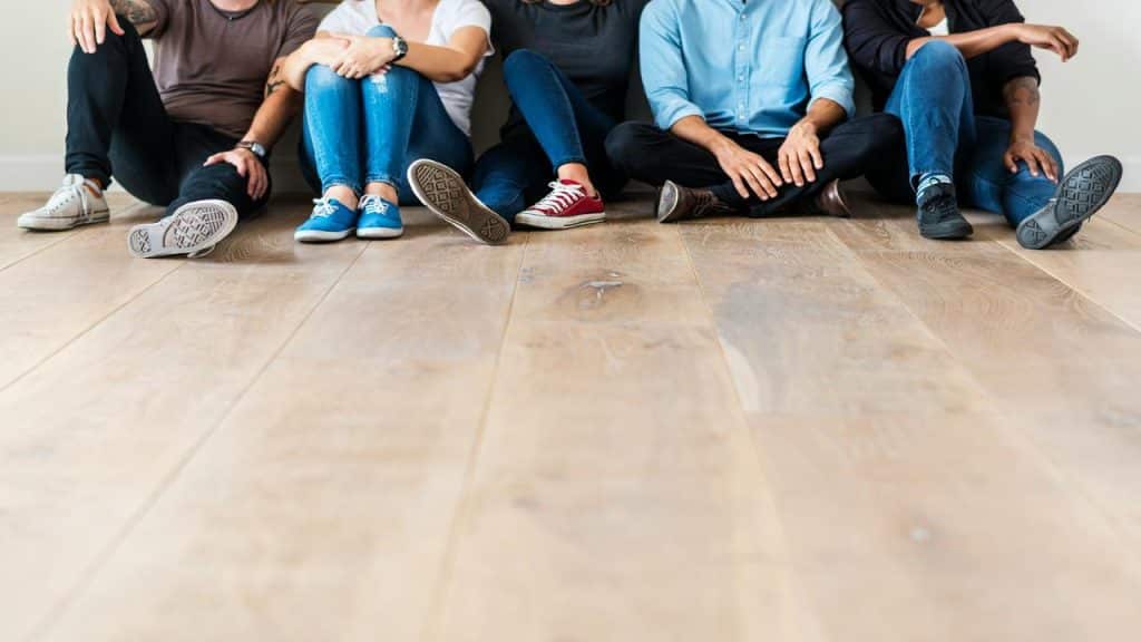 A group of six people sits casually on the floor against a wall, showing only their legs and feet.