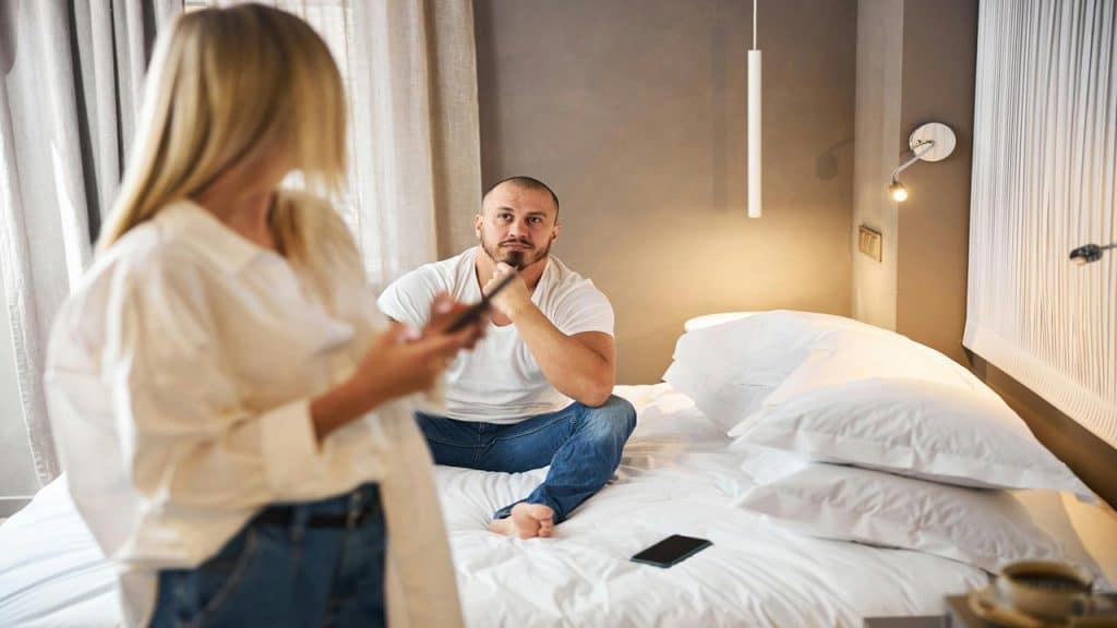 A man sits on a bed looking intently at a woman holding a phone, suggesting tension.