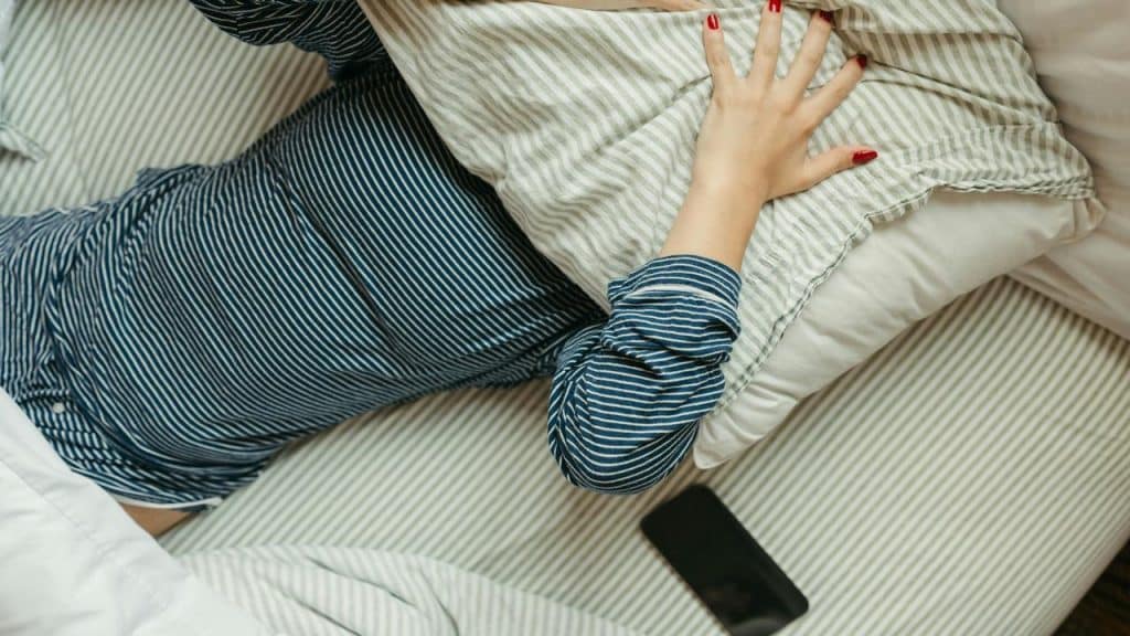 A person in striped pajamas lying in bed with a pillow over their face and a phone nearby.