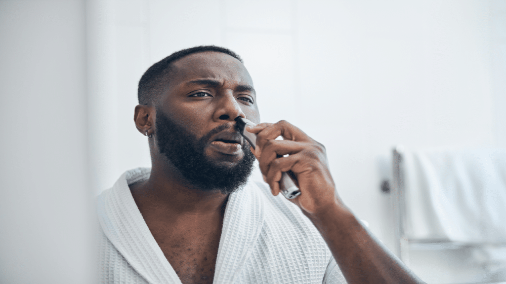 A man in a white bathrobe using a nose hair trimmer in a bright bathroom.
