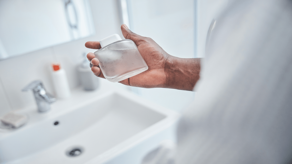 A close-up of a man holding a frosted glass bottle of cologne in a modern bathroom.