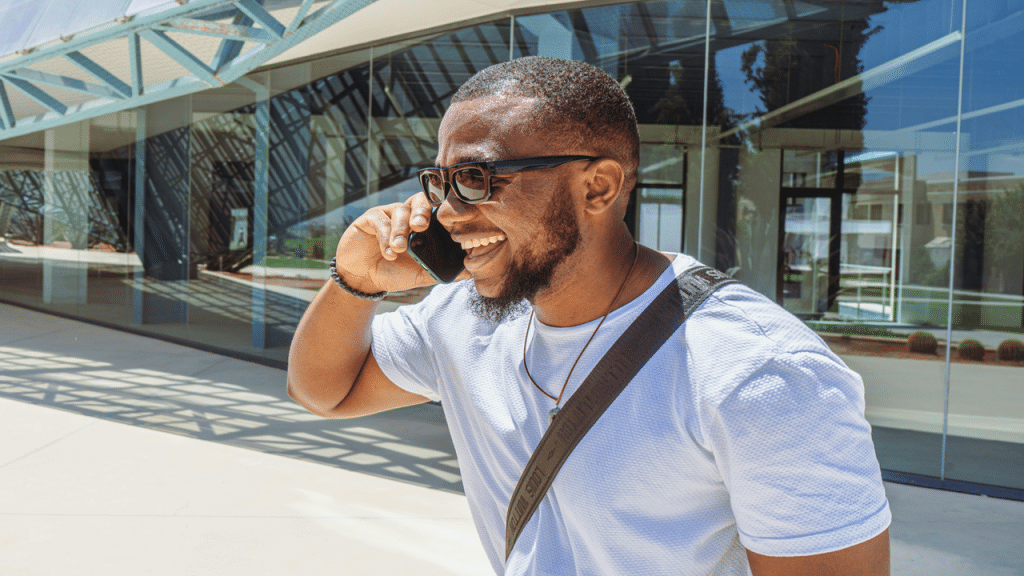 A smiling man in sunglasses talks on the phone outside a modern glass building.
