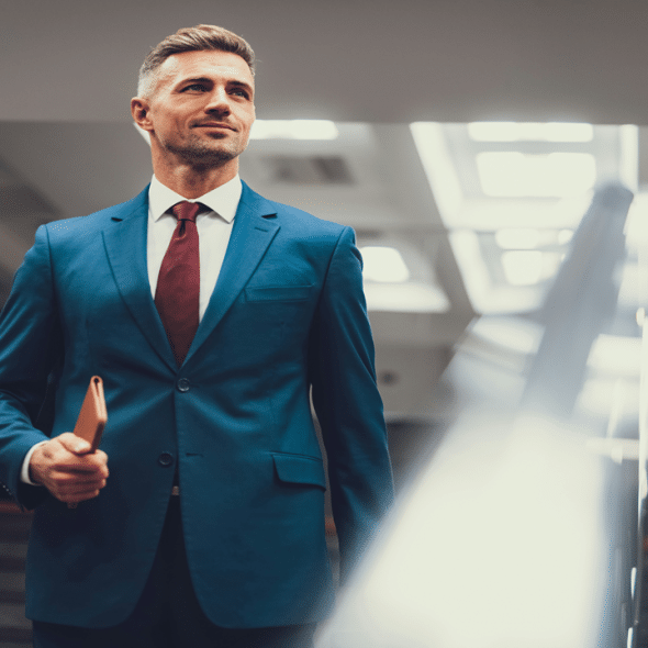 A confident businessman in a blue suit stands on a stair holding a notebook.