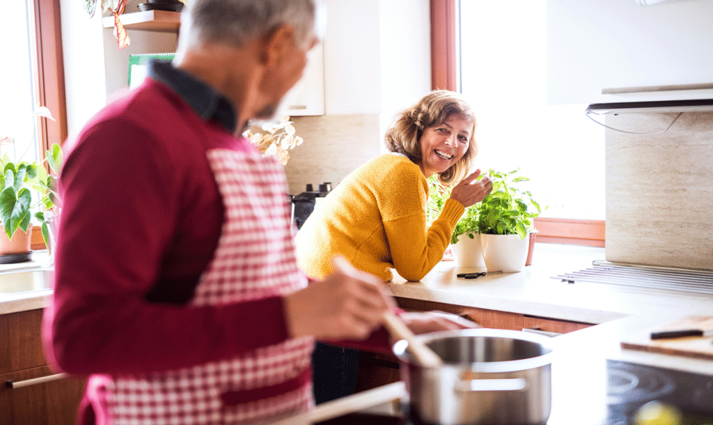 A cheerful older couple shares a moment while cooking together in a bright kitchen.